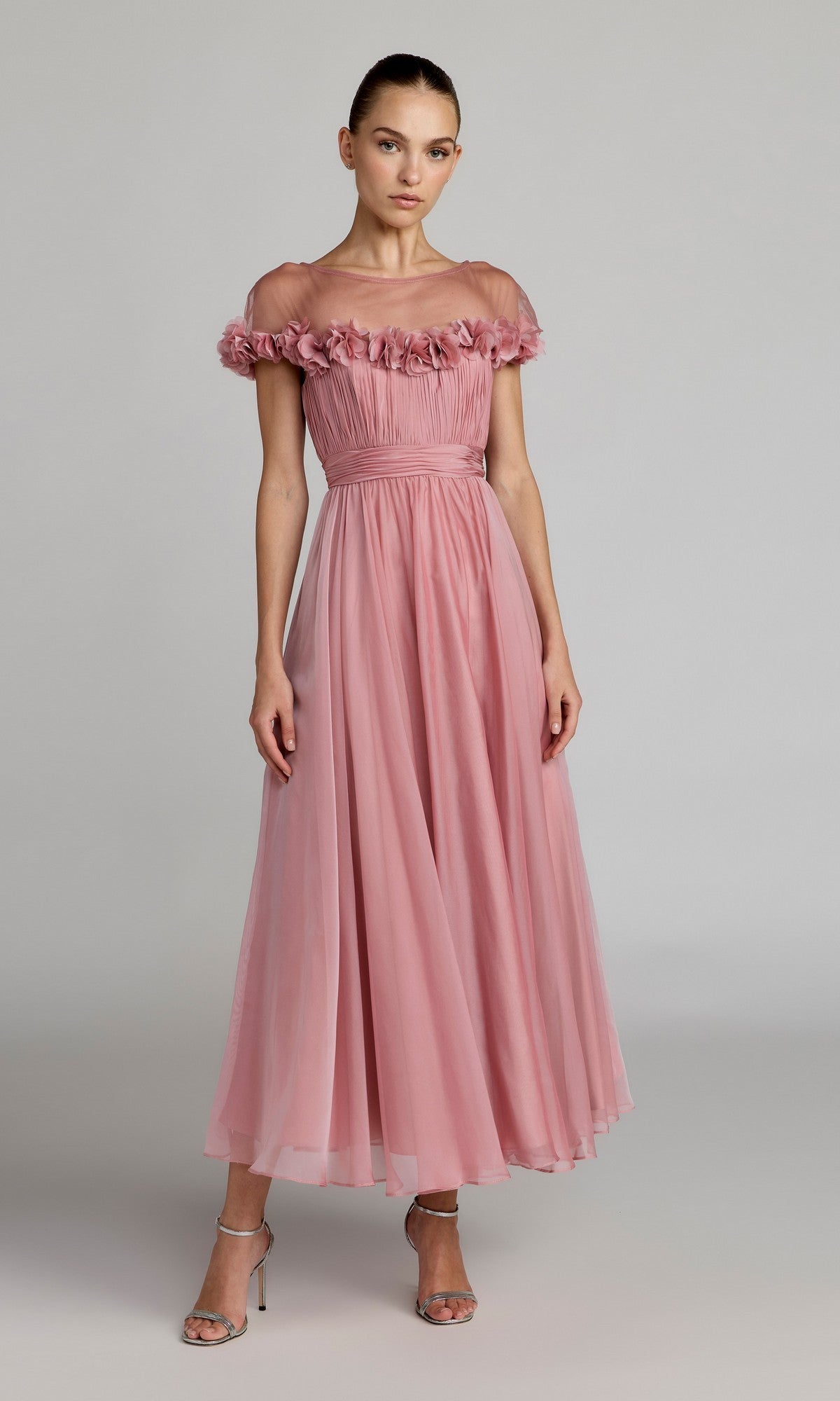 A woman wearing the Mac Duggal 68966 Tea-Length Party Dress in rose gold, featuring an off-the-shoulder ruffled neckline and ankle-length hem, stands in silver high-heeled sandals against a plain light background.
